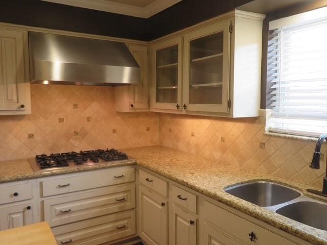 kitchen featuring stainless steel gas cooktop, backsplash, glass insert cabinets, a sink, and wall chimney exhaust hood