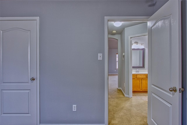 hall featuring arched walkways, baseboards, a sink, and light colored carpet