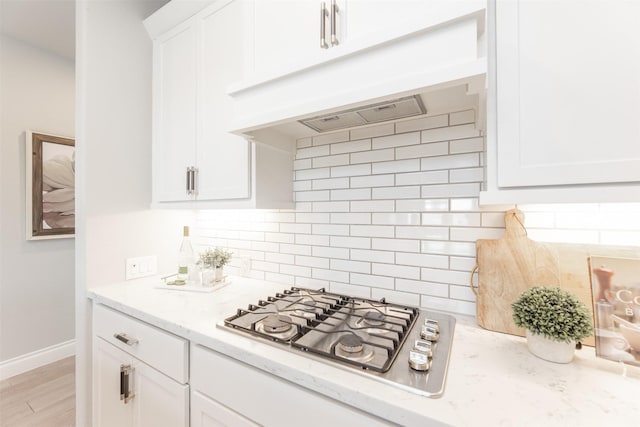 kitchen featuring light stone counters, range hood, stainless steel gas stovetop, backsplash, and white cabinets