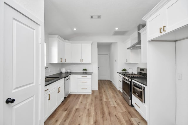 kitchen featuring dark countertops, appliances with stainless steel finishes, white cabinets, a sink, and wall chimney range hood