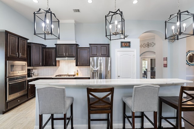 kitchen featuring arched walkways, dark brown cabinetry, under cabinet range hood, stainless steel appliances, and tasteful backsplash