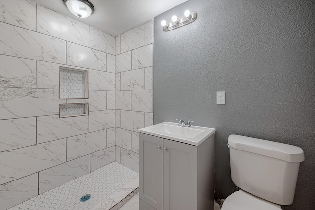 bathroom featuring vanity, toilet, a textured wall, and a tile shower