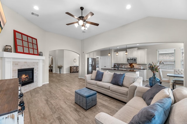 living room with visible vents, a brick fireplace, light wood-type flooring, arched walkways, and a ceiling fan