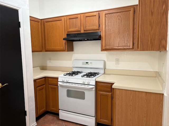 kitchen with dark tile patterned flooring, under cabinet range hood, gas range gas stove, brown cabinetry, and light countertops