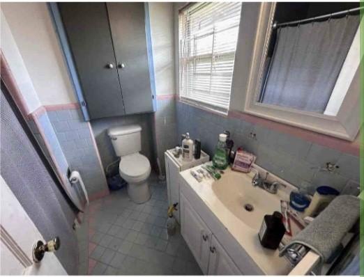 full bath with tile patterned flooring, a wainscoted wall, toilet, vanity, and tile walls