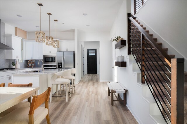 kitchen featuring tasteful backsplash, wall chimney range hood, light wood-style floors, stainless steel appliances, and a kitchen island with sink