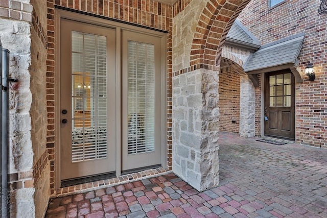 view of exterior entry featuring french doors, brick siding, and roof with shingles