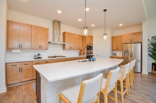 kitchen featuring light wood-type flooring, a kitchen island with sink, a sink, appliances with stainless steel finishes, and wall chimney range hood