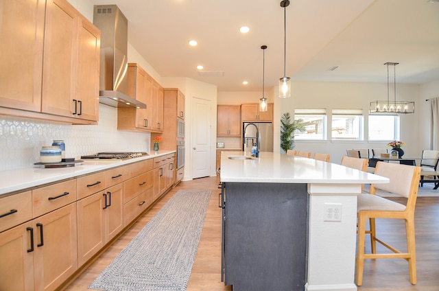 kitchen with light brown cabinetry, appliances with stainless steel finishes, wall chimney exhaust hood, and a kitchen bar