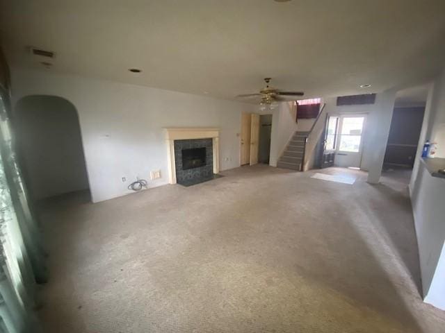 unfurnished living room featuring a tiled fireplace, carpet floors, arched walkways, ceiling fan, and stairs