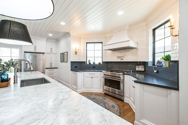 kitchen with high quality appliances, decorative backsplash, custom exhaust hood, white cabinetry, and a sink