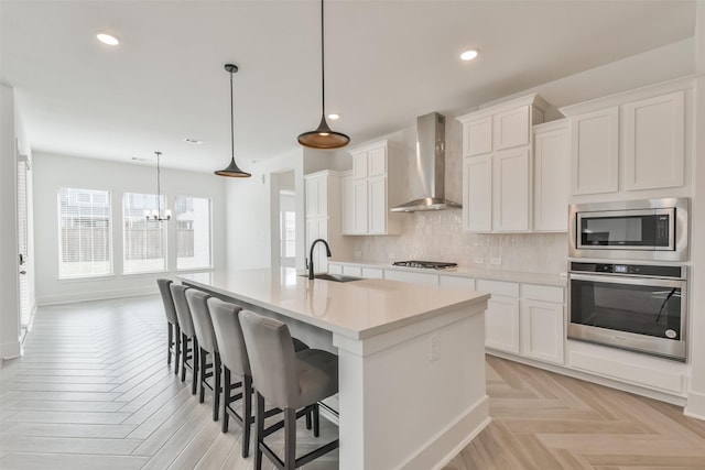 kitchen featuring an island with sink, a sink, backsplash, appliances with stainless steel finishes, and wall chimney range hood