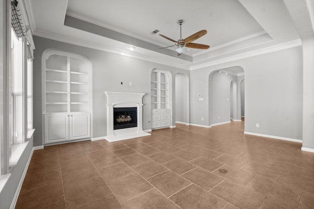 unfurnished living room with ceiling fan, a tray ceiling, built in shelves, and a fireplace with raised hearth