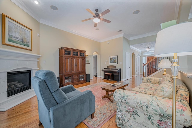 living room with light wood-type flooring, ornamental molding, a ceiling fan, a glass covered fireplace, and arched walkways