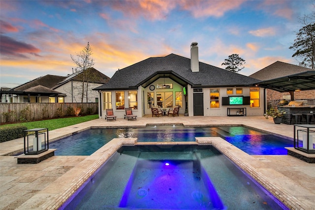 pool at dusk featuring outdoor dry bar, an exterior structure, fence, a fireplace, and a patio area