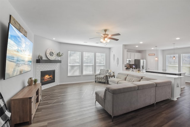 living room featuring a ceiling fan, baseboards, recessed lighting, dark wood-style flooring, and a brick fireplace