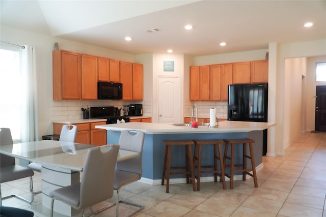 kitchen with backsplash, a breakfast bar, an island with sink, light countertops, and black appliances