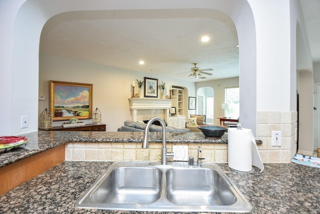 kitchen with a ceiling fan, dark stone counters, a fireplace, arched walkways, and a sink