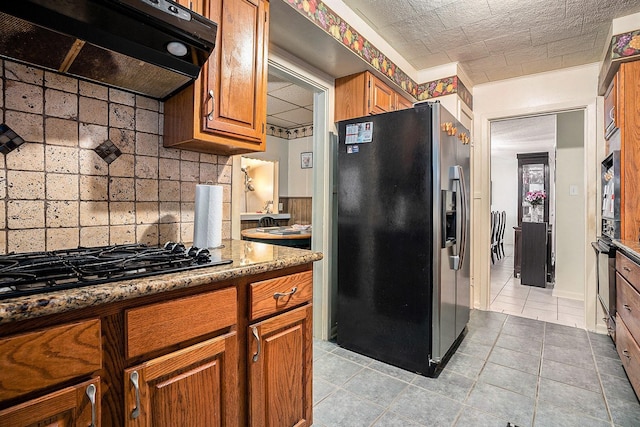 kitchen featuring black gas stovetop, under cabinet range hood, brown cabinetry, stainless steel fridge with ice dispenser, and decorative backsplash