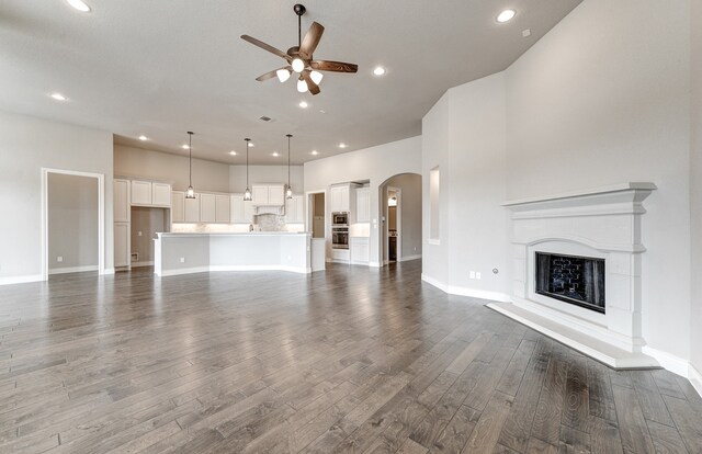 unfurnished living room with ceiling fan, arched walkways, a fireplace with raised hearth, and dark wood-style flooring