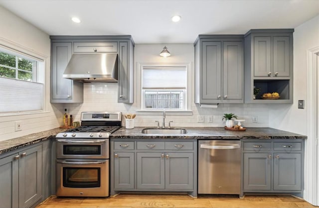 kitchen featuring ventilation hood, dark stone countertops, gray cabinets, appliances with stainless steel finishes, and a sink