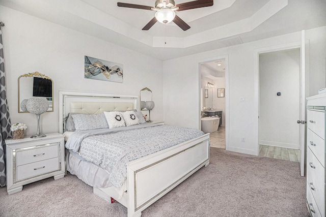 bedroom featuring a tray ceiling, light colored carpet, ensuite bath, and a ceiling fan