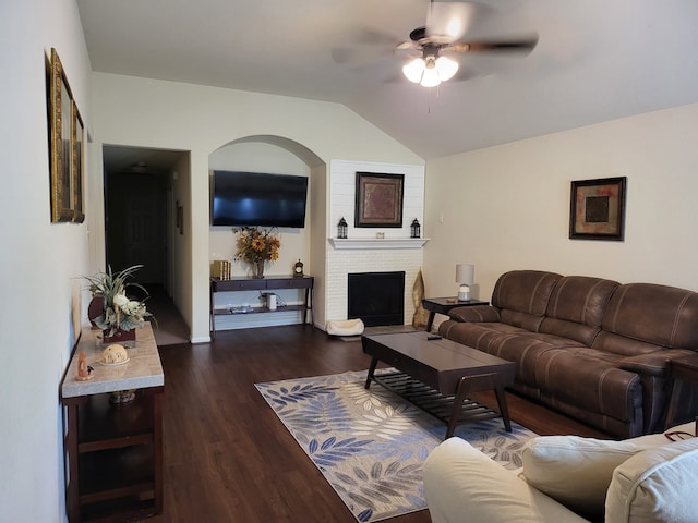 living room with a ceiling fan, vaulted ceiling, a brick fireplace, and dark wood-style floors