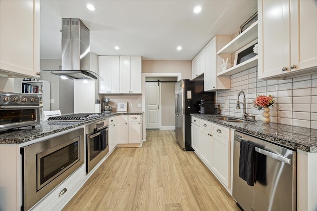 kitchen with wall chimney range hood, light wood-type flooring, a barn door, appliances with stainless steel finishes, and open shelves