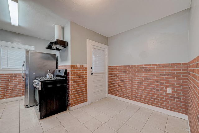 kitchen featuring light tile patterned flooring, black gas range, brick wall, and a wainscoted wall