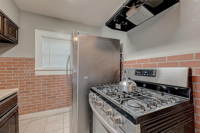 kitchen with dark brown cabinets, brick wall, ventilation hood, light tile patterned floors, and appliances with stainless steel finishes