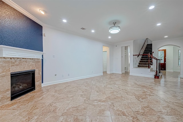 unfurnished living room with arched walkways, crown molding, a tiled fireplace, and stairs