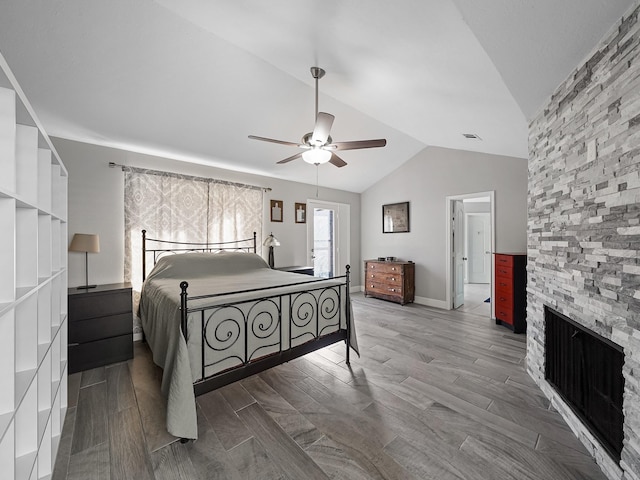 bedroom with ceiling fan, baseboards, wood finish floors, lofted ceiling, and a stone fireplace