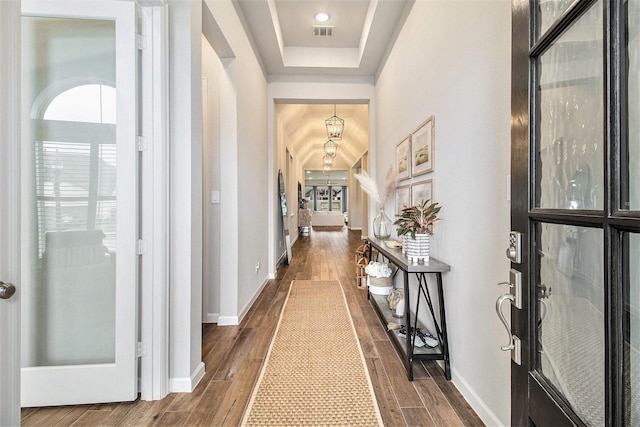 hall with visible vents, a tray ceiling, baseboards, and dark wood-style flooring