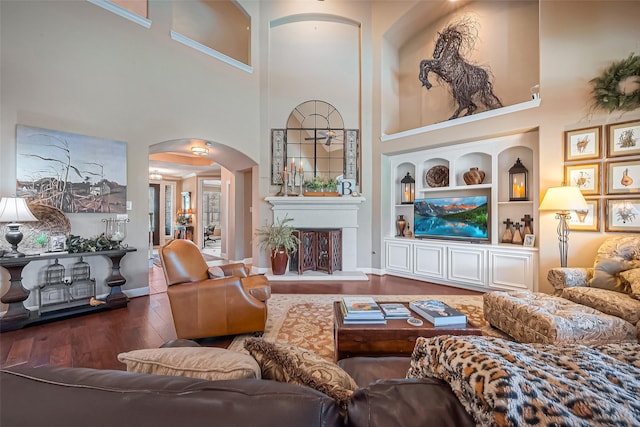 living room featuring built in features, wood finished floors, baseboards, a fireplace with raised hearth, and arched walkways