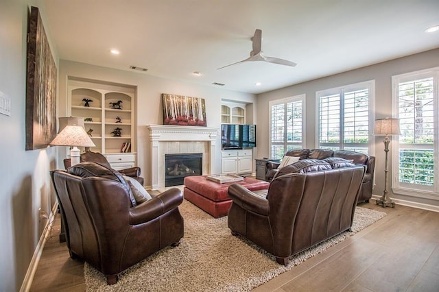 living room with built in shelves, baseboards, a tile fireplace, wood finished floors, and a ceiling fan