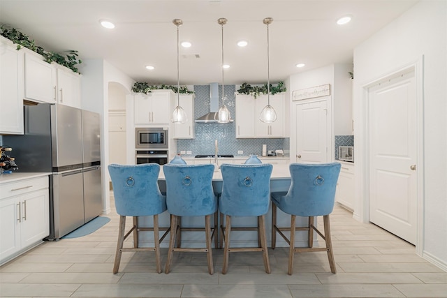 kitchen with a breakfast bar, an island with sink, stainless steel appliances, white cabinetry, and wall chimney range hood