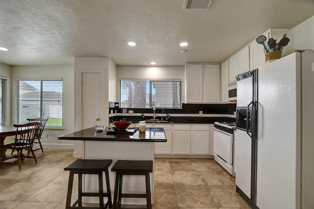 kitchen with white appliances, a kitchen island, a sink, white cabinetry, and dark countertops