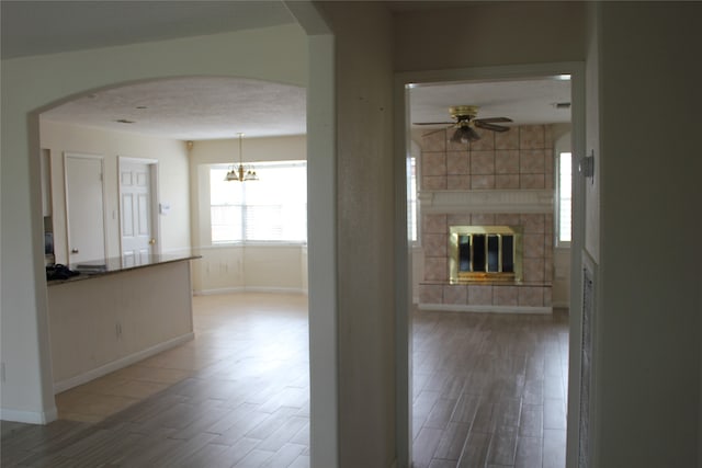 unfurnished living room featuring wood finished floors, baseboards, arched walkways, a tiled fireplace, and ceiling fan with notable chandelier