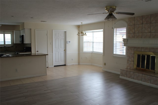 unfurnished living room featuring baseboards, a tiled fireplace, ceiling fan with notable chandelier, light wood-style flooring, and a textured ceiling