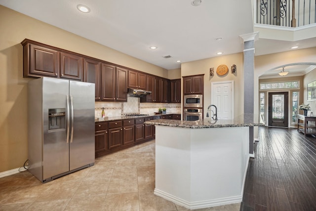 kitchen with dark stone counters, arched walkways, stainless steel appliances, under cabinet range hood, and backsplash