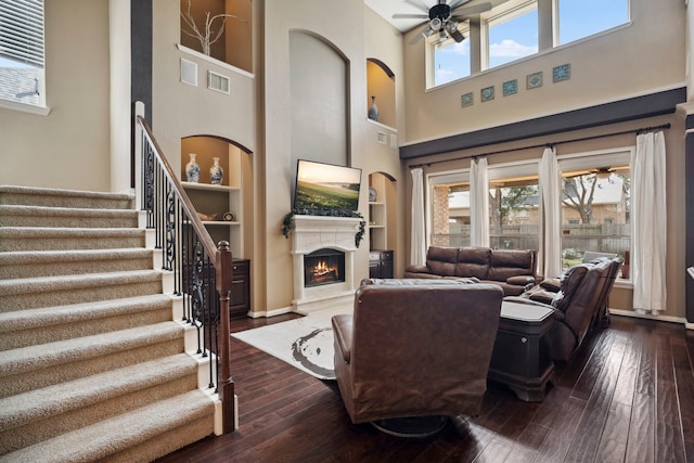 living area with stairs, a lit fireplace, ceiling fan, and dark wood-style flooring