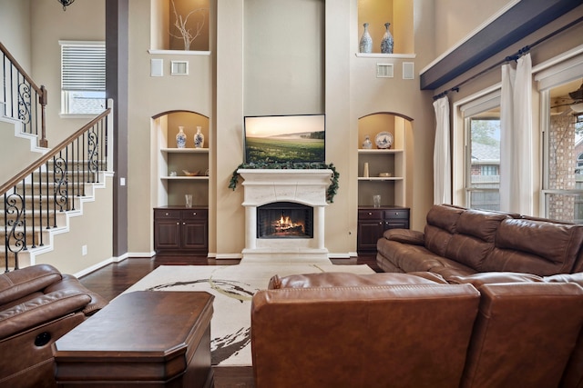 living room featuring stairway, built in features, baseboards, dark wood-style flooring, and a warm lit fireplace