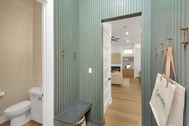 bathroom with wood finished floors, wallpapered walls, a fireplace, ceiling fan, and toilet