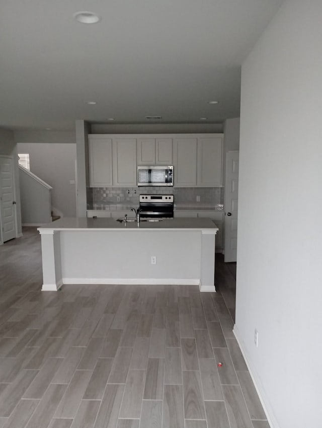 kitchen with light wood-style flooring, an island with sink, backsplash, appliances with stainless steel finishes, and light countertops