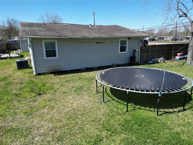 back of property with fence, roof with shingles, central air condition unit, a trampoline, and a lawn