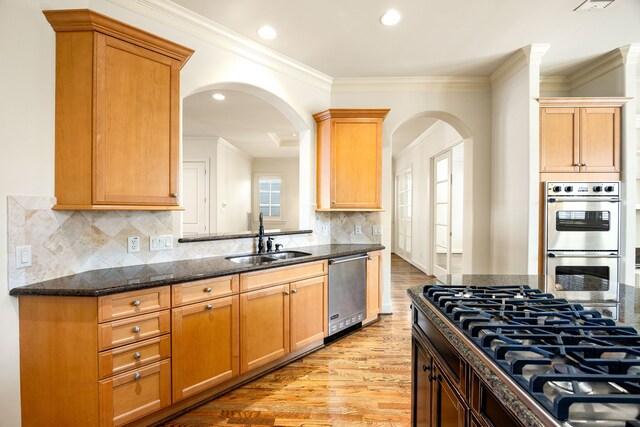 kitchen with ornamental molding, dark stone countertops, appliances with stainless steel finishes, light wood-style floors, and a sink