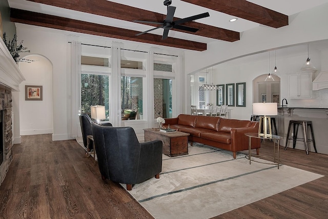 living room with beam ceiling, arched walkways, a stone fireplace, and dark wood-style flooring