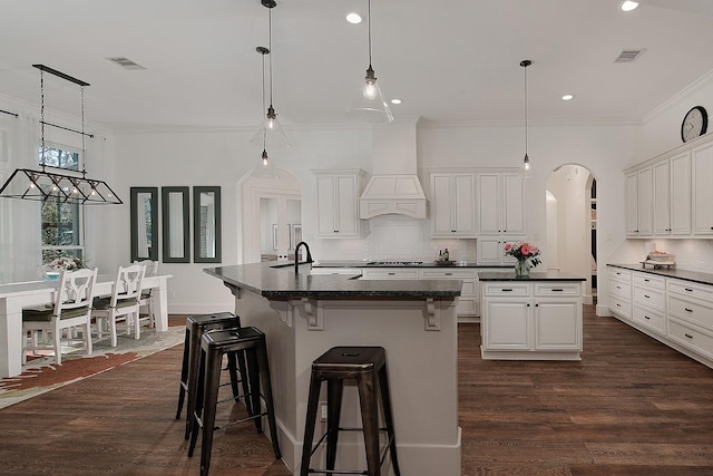 kitchen featuring dark countertops, visible vents, an island with sink, arched walkways, and custom exhaust hood