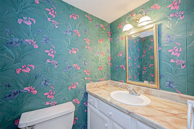 half bath with toilet, wallpapered walls, a textured ceiling, and vanity