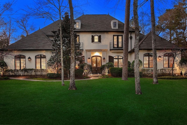 french country style house featuring a front yard, a balcony, a shingled roof, stucco siding, and french doors